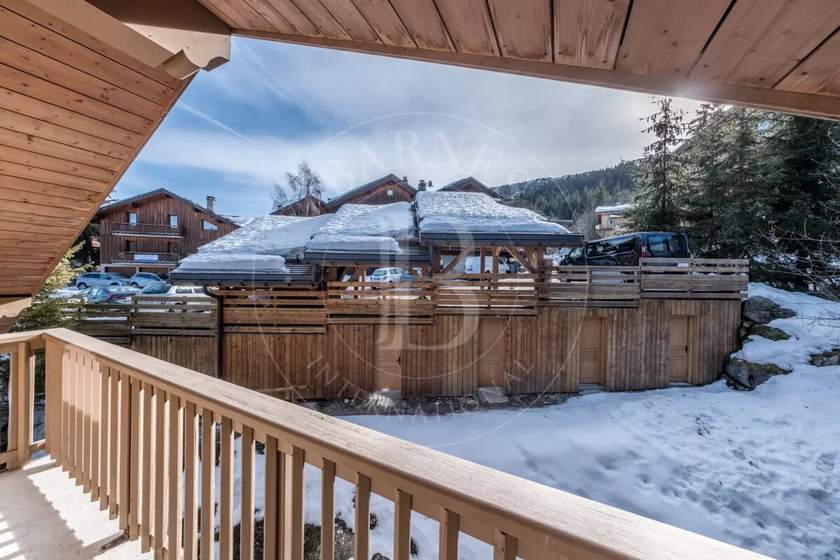 View from a wooden balcony overlooking a snowy mountain village. Traditional wooden chalets with snow-covered roofs, parked cars, and pine trees fill the winter landscape.