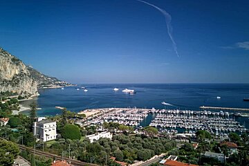 Stunning aerial view of a bustling coastal marina packed with yachts, clear blue sea, dramatic cliffs, and hillside buildings.