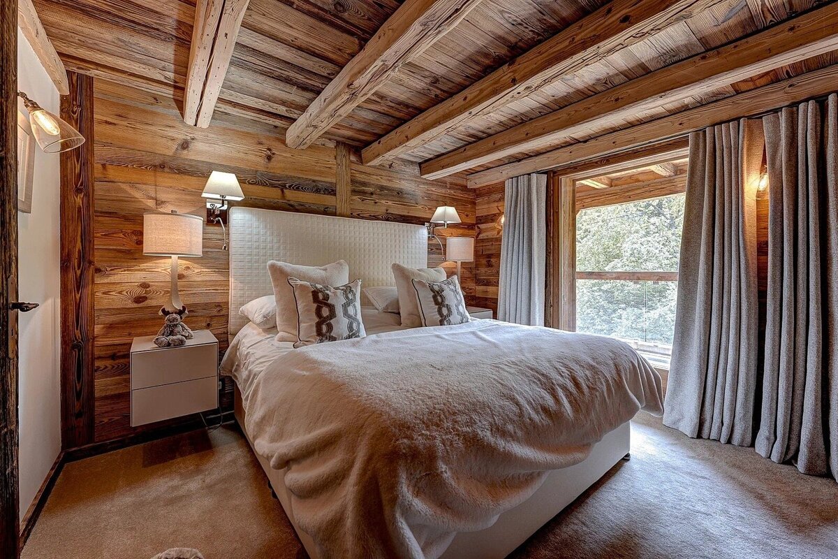 Warm, rustic bedroom featuring exposed wood walls/ceiling, a plush white bed, and a large window with a scenic view.