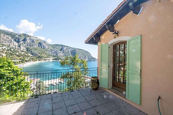 A balcony offers a scenic view of a vibrant turquoise bay, green mountains, and a coastal town. A peach building with open green shutters stands on the right.