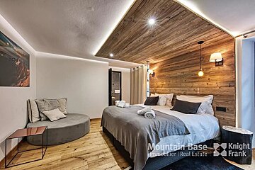 A cozy, modern bedroom with extensive wood paneling, a large bed, and a circular chaise lounge. Accent lighting highlights the warm, inviting ambiance.