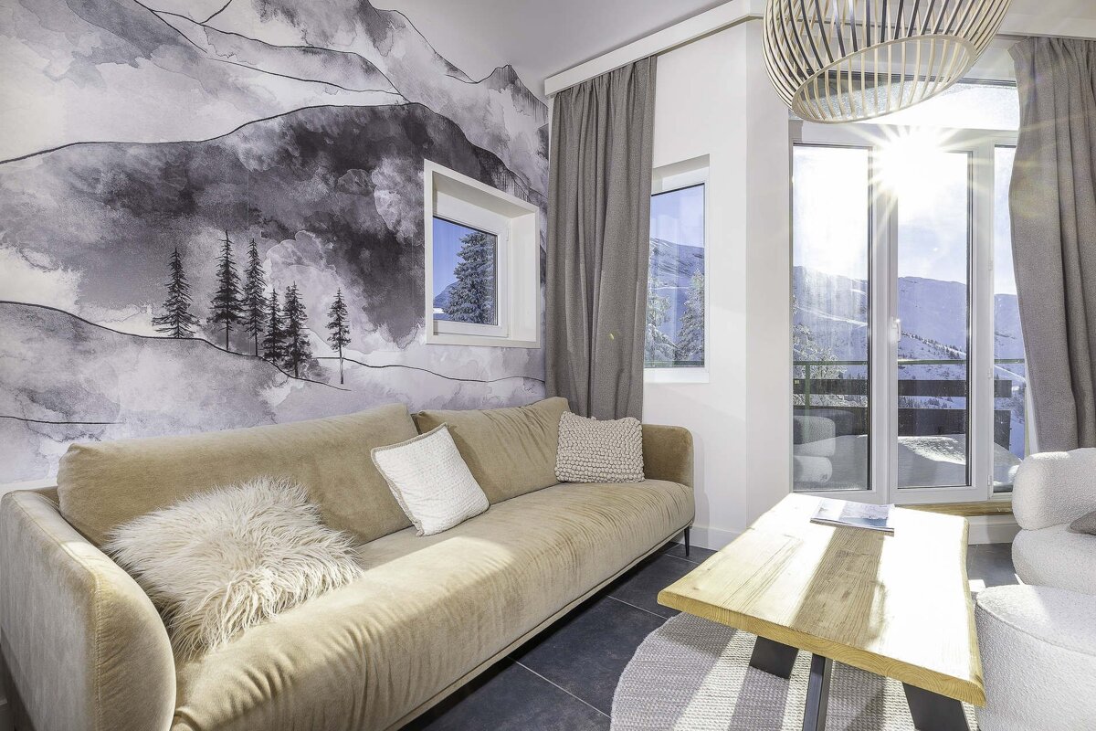 A cozy, modern room features a mountain mural, a long beige sofa, and a window offering a bright, sunlit view of snowy mountains.