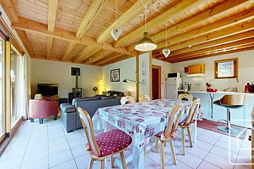 Cozy open-concept room with wood-beamed ceiling. Dining area, living space with wood stove & TV, and a kitchenette with bar stools.