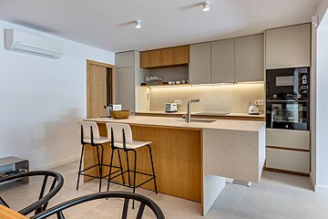 Modern kitchen with a light wood island, bar stools, light gray cabinets, built-in appliances, and illuminated backsplash.
