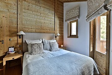 A cozy, rustic bedroom with warm wood-paneled walls, a bed with furry pillows, patterned blinds on windows, and a door leading outside.