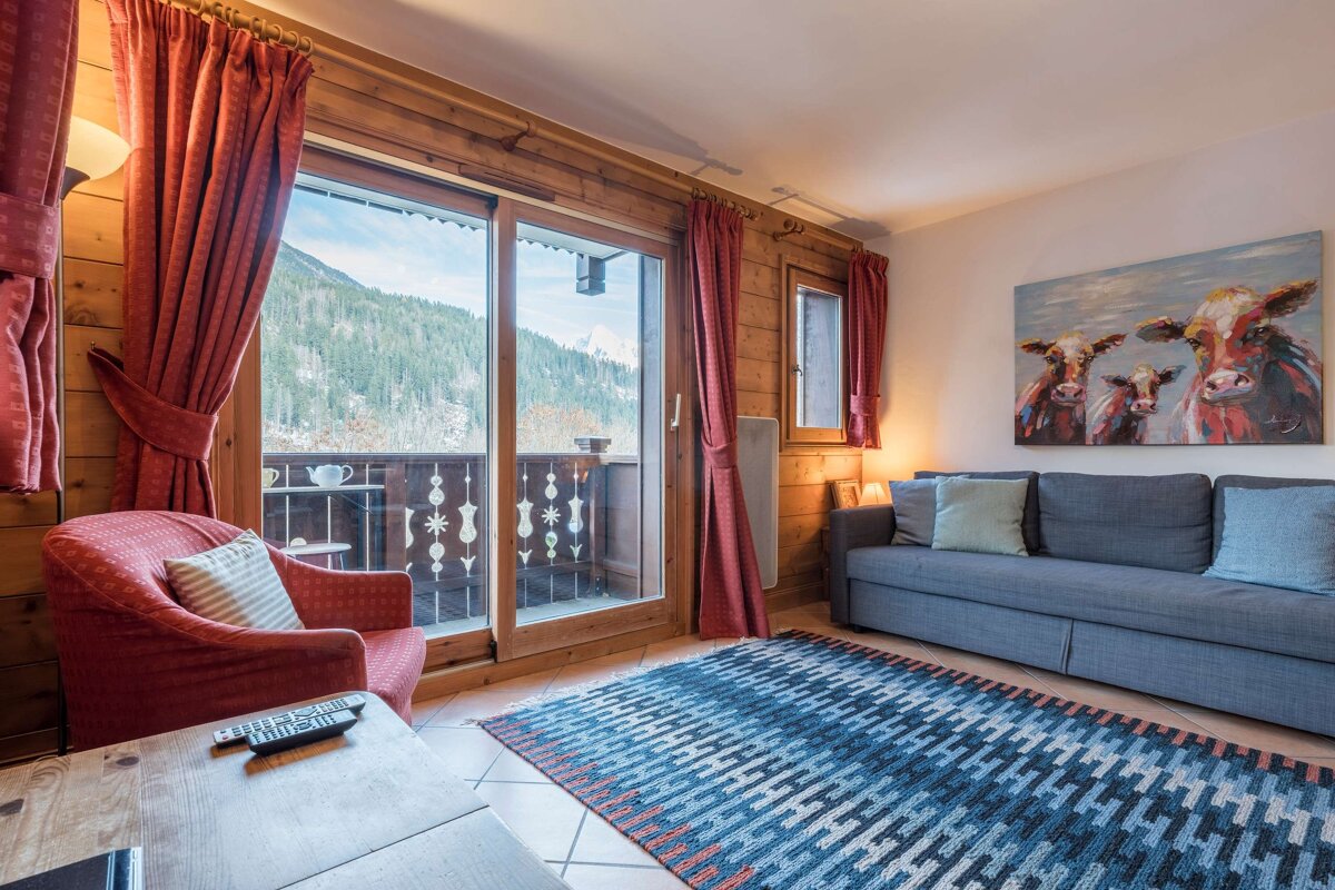 Cozy cabin living room with wood-paneled walls, a blue sofa, red curtains, and sliding glass doors opening to scenic mountain views.