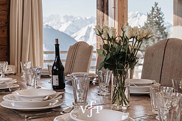 Elegantly set dining table with white roses and a wine bottle in a cozy chalet overlooking snowy mountains.