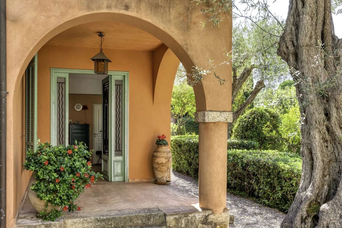 Tuscan-style arched entrance with a mint green door, hanging lantern, and potted flowers, leading to a serene garden with an ancient olive tree.