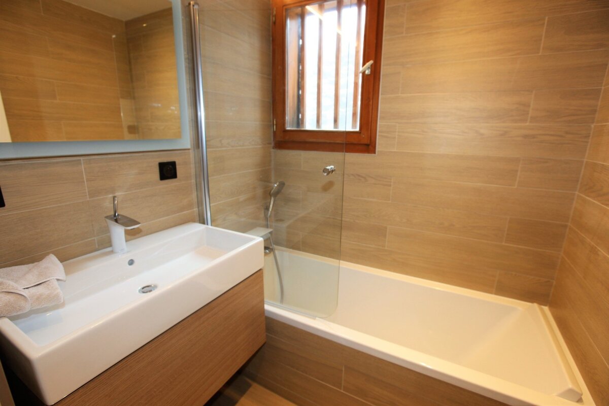 Modern bathroom featuring wood-look tile walls, a white vanity sink, a bathtub with a glass partition, and a small window.