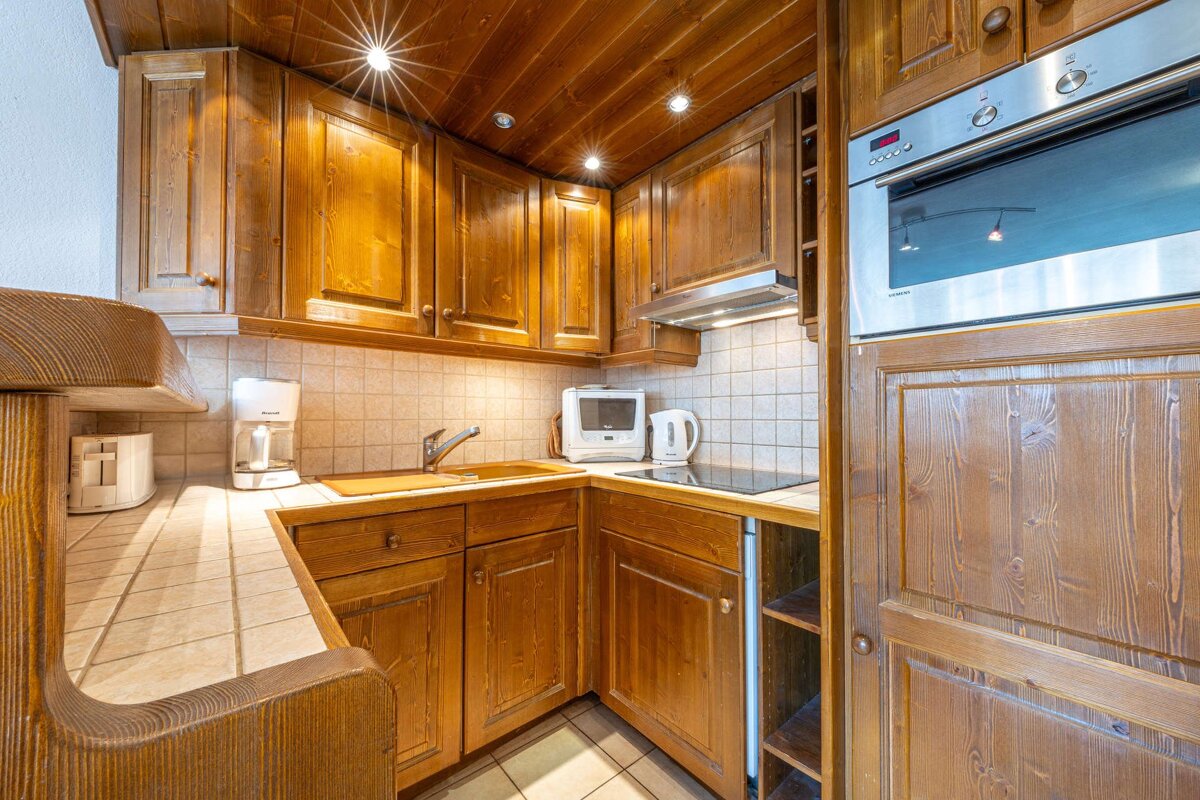 A compact kitchen with light brown wooden cabinets, tiled counters, and recessed ceiling lights. It features modern appliances like an oven, stovetop, coffee maker, and toaster.