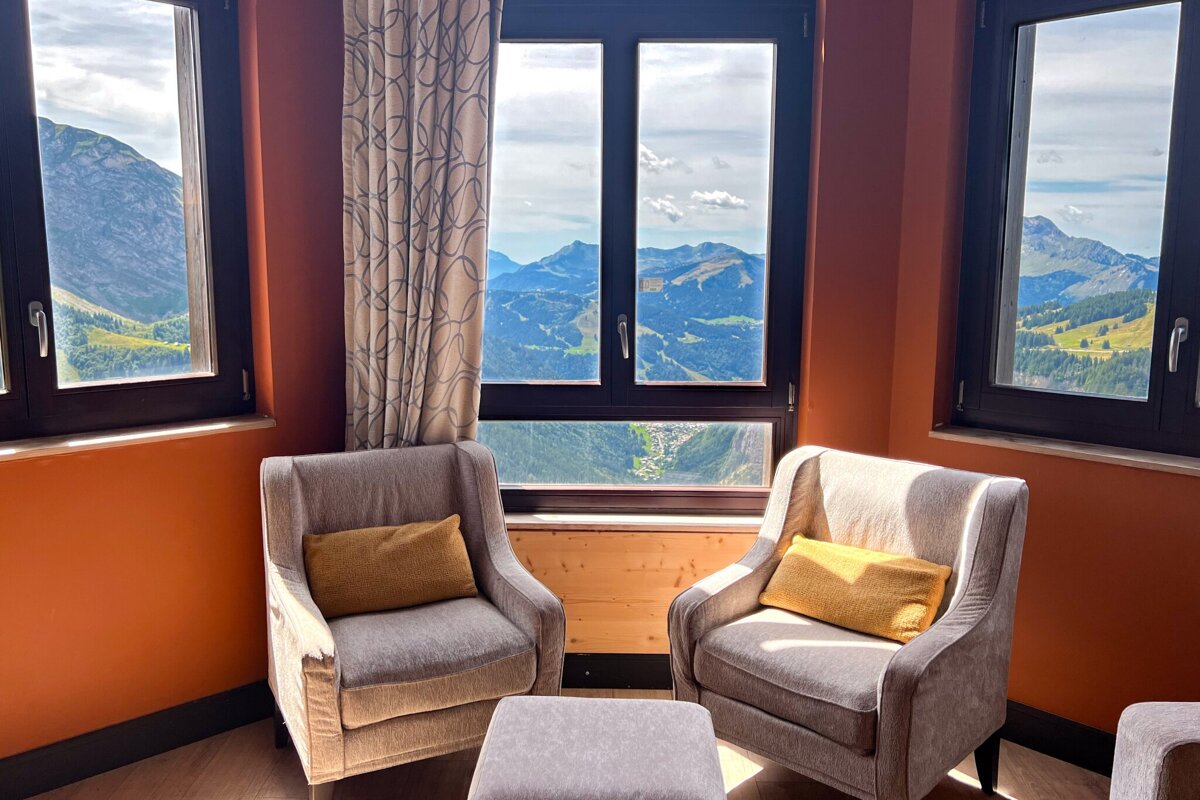 An inviting room with two armchairs, an ottoman, and warm orange walls, offering panoramic mountain views through multiple windows.