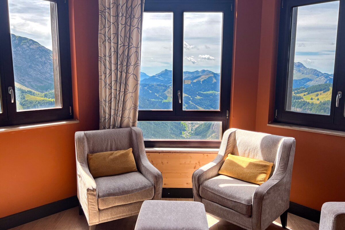 Cozy armchairs face large windows offering a scenic, panoramic view of sunny green mountains.