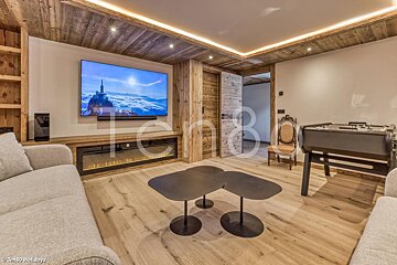 A cozy, modern chalet living room features a large TV above an electric fireplace, warm recessed lighting, wooden walls/ceiling, a foosball table, and a plush sofa.