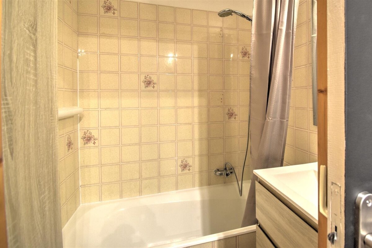 A bathroom featuring a bathtub with beige-tiled walls, a showerhead, curtains, and a sink vanity on the right.