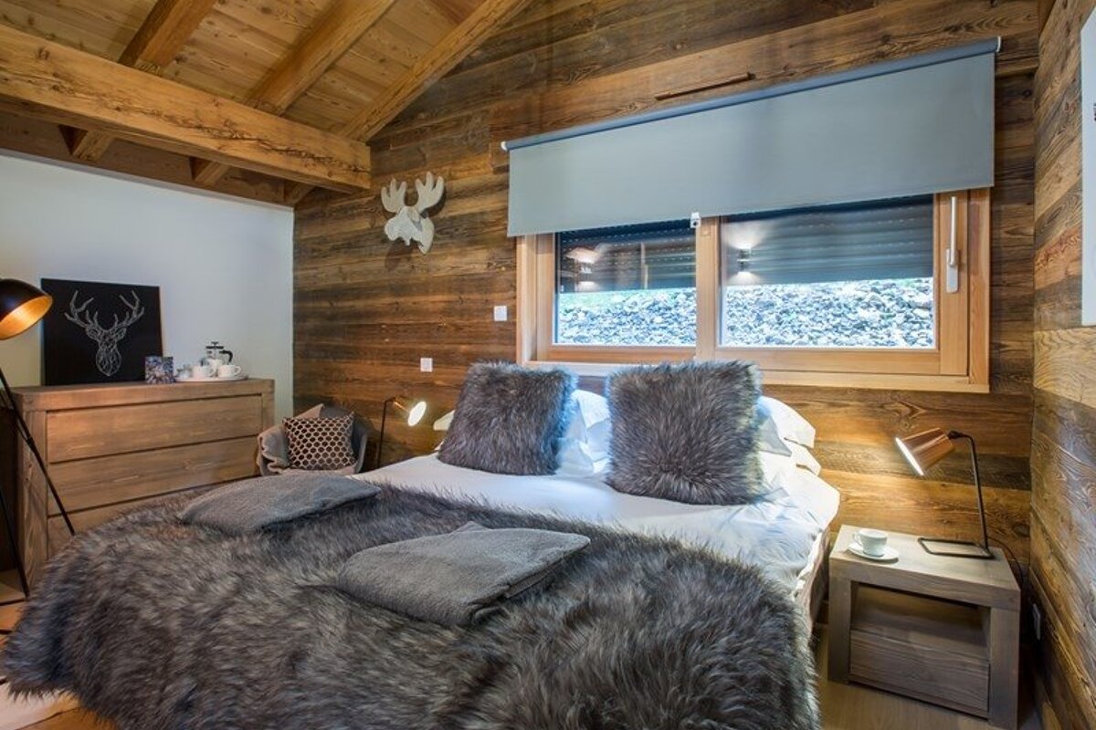 Cozy rustic chalet bedroom featuring wooden walls, a bed with fluffy grey faux-fur blankets, and warm ambient lighting.