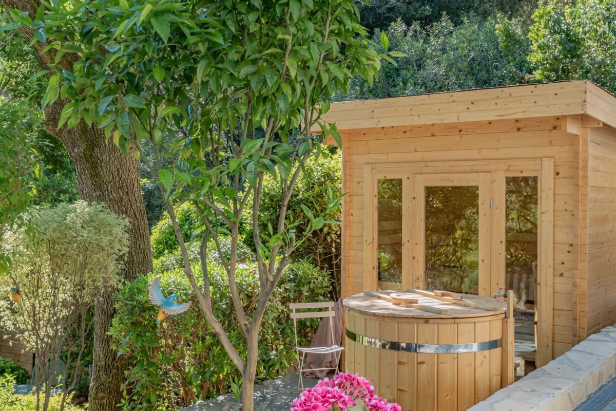 A cozy wooden sauna cabin and matching hot tub are nestled among lush green trees and foliage in a vibrant outdoor garden setting.