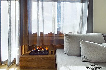 A modern, cozy interior featuring bright windows with sheer curtains, a wooden base with a faux fireplace, and a plush grey sofa.