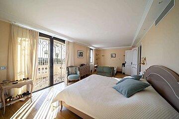 A bright, sunlit bedroom with a large bed, blue pillows, and a balcony with an ornate railing. Sunlight streams through glass doors onto the wooden floor.