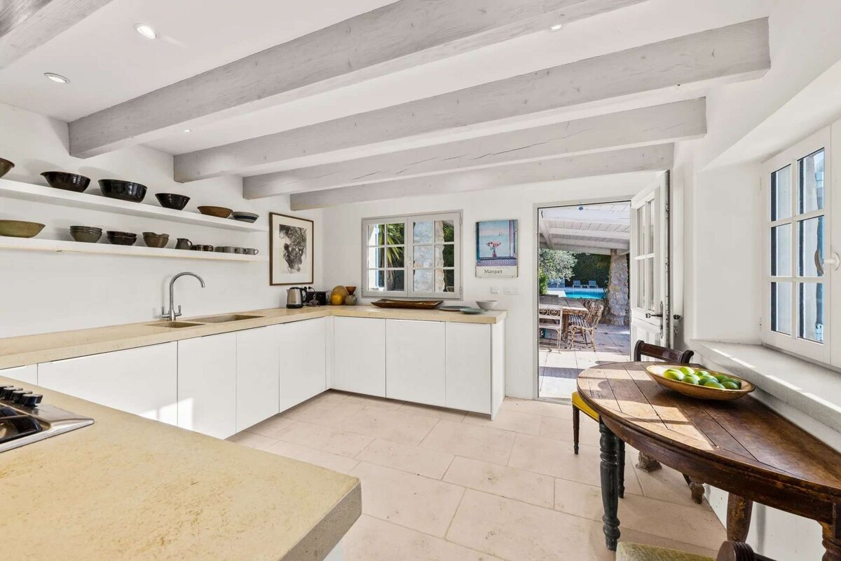 A bright, Mediterranean-style kitchen with white cabinets, stone counters, and wooden beams. A door opens to a patio with a pool, and a small dining table sits nearby.