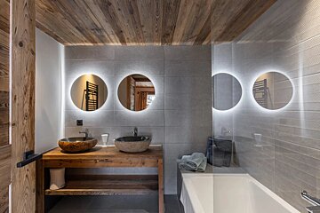 Modern rustic bathroom featuring wood elements, grey tiled walls, two bowl sinks under backlit circular mirrors, and a sleek bathtub.