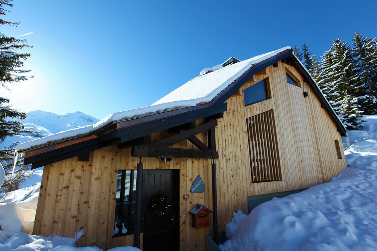 A modern wooden chalet surrounded by deep snow and pine trees under a clear blue sky.