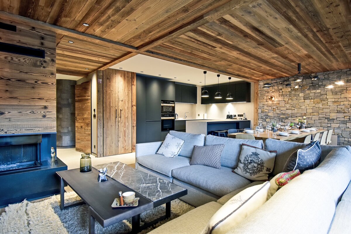 A cozy open-plan living space with a wooden ceiling, stone wall, modern dark kitchen, large gray sofa, and dining area, creating a rustic-chic vibe.