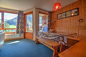 Rustic dining room with wood-paneled walls, wooden benches, blue carpet, and large windows looking out at green mountains.