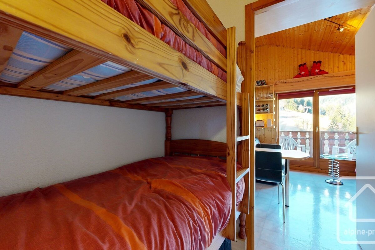 A cozy room features a wooden bunk bed with an orange duvet in the foreground. Beyond it, a living area with a balcony overlooks a snowy landscape.