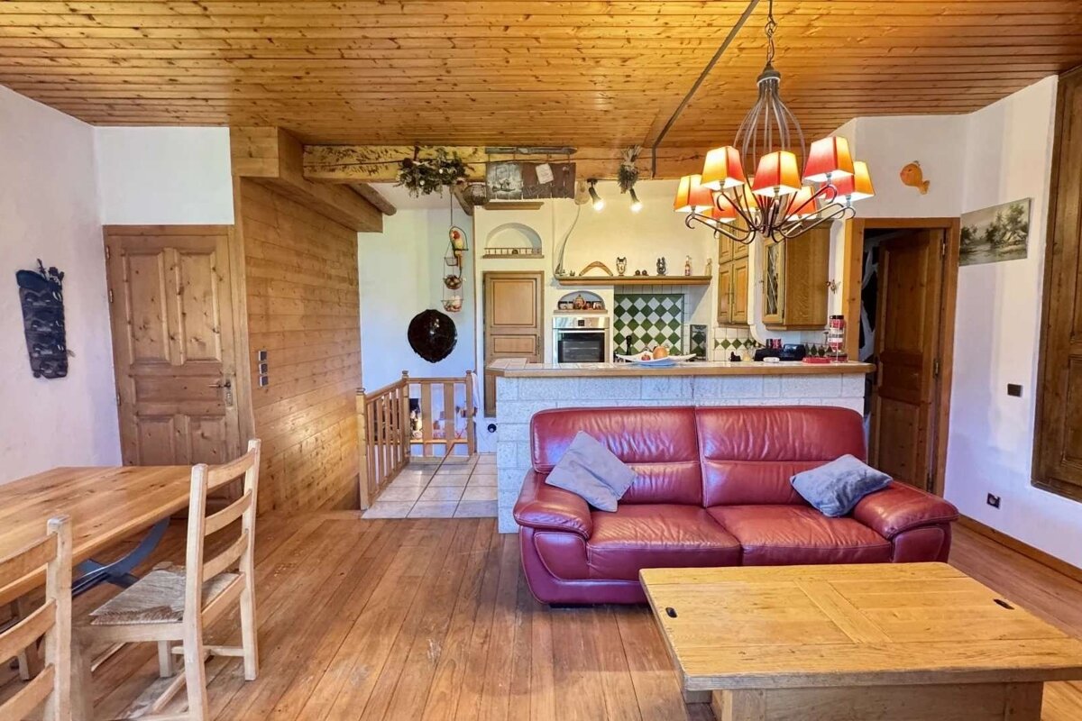 Cozy, rustic open-plan living room with wood ceilings/floors, a red leather sofa, wooden dining table, and a kitchen with a bar.