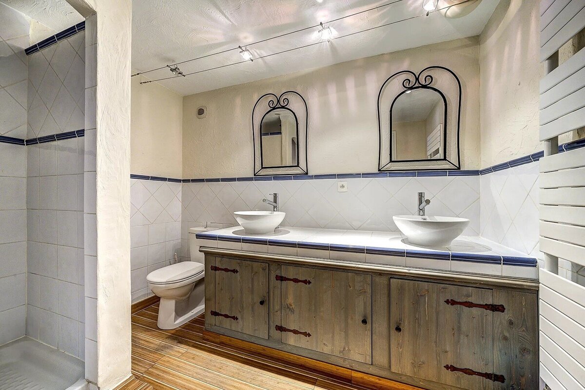 A rustic bathroom with a double wooden vanity, two white vessel sinks, and ornate black mirrors. Features a shower, toilet, and blue-accented white tiled walls.