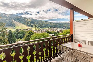 A charming balcony offers a view of a picturesque mountain village nestled in a lush valley, surrounded by forested slopes and a visible ski run under a cloudy sky.