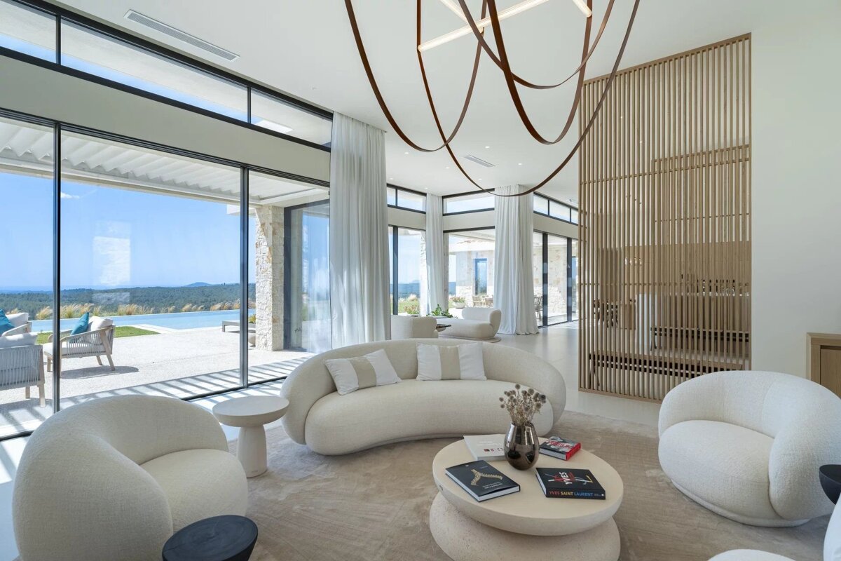 Chic, modern interior with white curved furniture, expansive windows showing a pool and natural vista, and a unique flowing ceiling light.