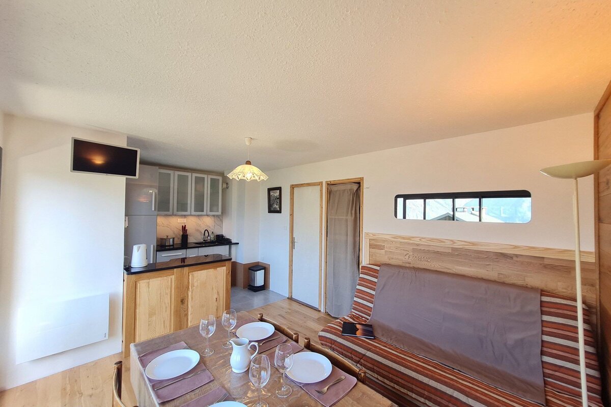 A bright, compact studio apartment with a set dining table, integrated kitchen, and striped sofa bed, featuring warm wood accents and modern details.