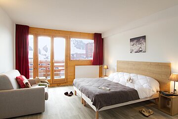 A warm, modern bedroom in a ski chalet with a large window framing a snowy mountain view. Features a bed, sofa, wooden accents, and red curtains.