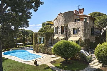 A luxurious stone villa with a curved facade, lush green ivy, a swimming pool, and manicured gardens under a clear blue sky.