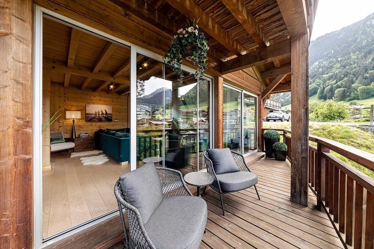 A wooden chalet balcony with comfy chairs overlooks a mountain vista. Glass doors reveal a cozy interior, all in warm wood tones.