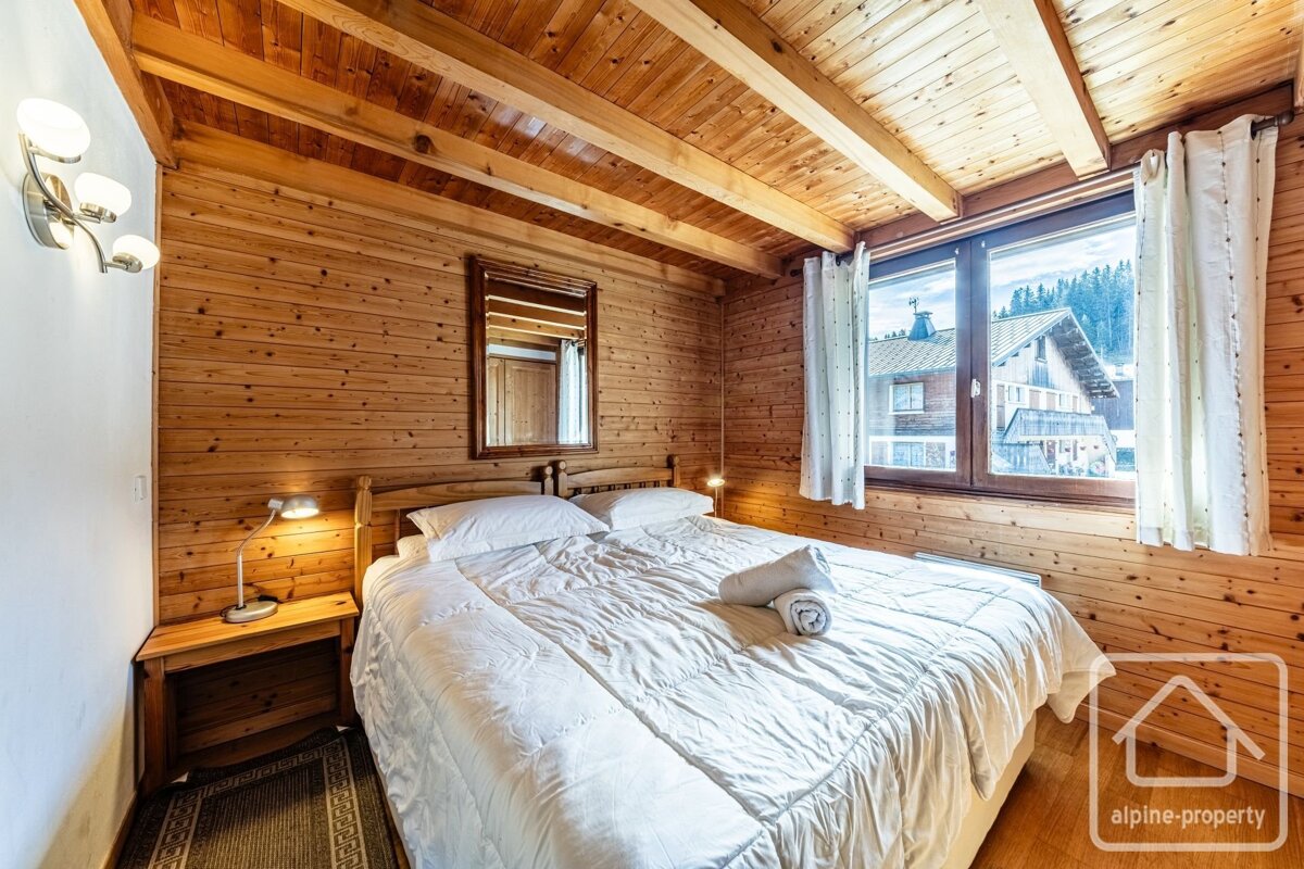 Cozy alpine bedroom with warm wooden walls and ceiling, a double bed, and a window showcasing a village and mountain view.