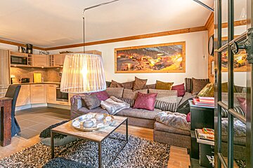 Cozy open-plan living/kitchen with a large L-shaped sofa, many pillows, a modern pendant light, and a prominent mountain village artwork.
