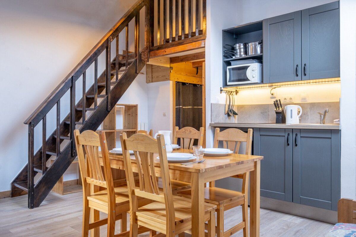 A cozy room featuring a wooden dining table, a modern gray kitchenette under bright lighting, and a dark wooden staircase ascending to a mezzanine level.