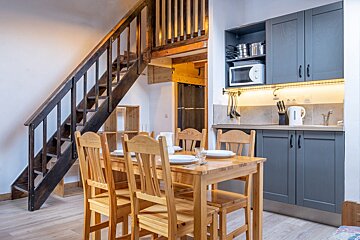 A cozy room featuring a wooden dining table, a modern gray kitchenette under bright lighting, and a dark wooden staircase ascending to a mezzanine level.