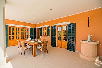 A bright, orange-walled outdoor patio features a set dining table for six, wooden doors with dark shutters, and a pitcher on a decorative well.