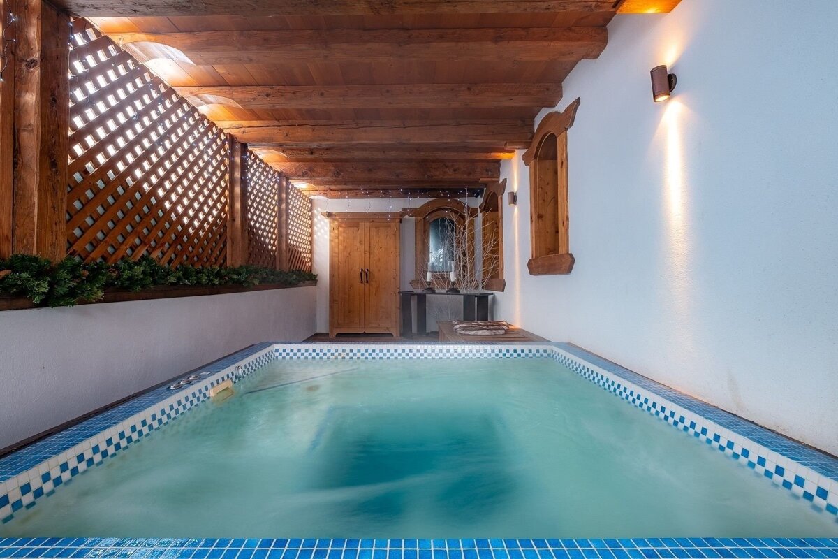 A cozy indoor pool area with blue tiled edges, a wooden lattice wall, exposed beams, and warm lighting creating a rustic ambiance.