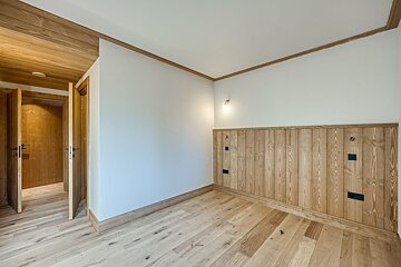 An empty room with light wood floors, white walls, and a wood-paneled accent wall. Two open wooden doors reveal a hallway, all trimmed in wood.