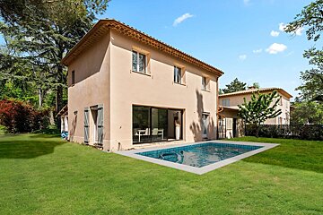 A two-story beige Mediterranean villa with a swimming pool set in a lush green lawn under a clear blue sky.