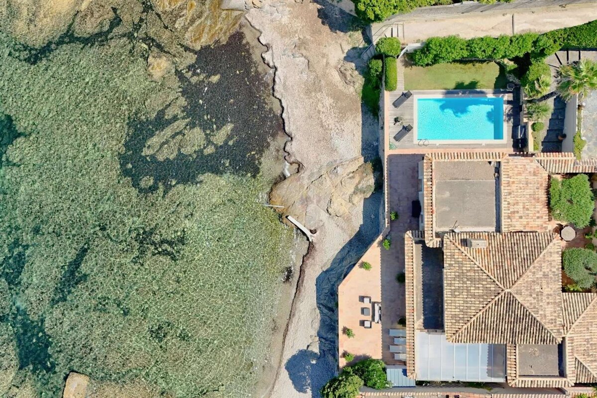 Aerial view of a coastal property featuring clear turquoise water, a rocky beach, a house with tiled roofs, and a bright blue swimming pool surrounded by greenery.