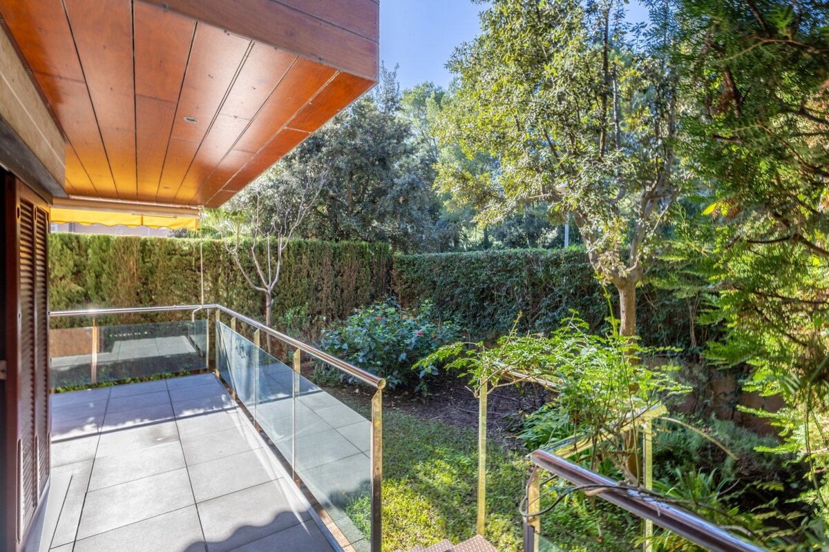 A sunny balcony with tiled floor, glass railing, and a wooden ceiling overlooks a lush green garden filled with trees and hedges.