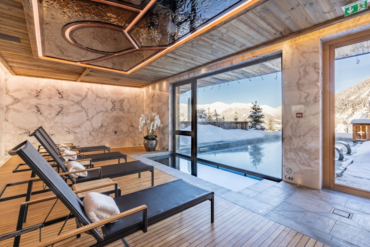 Luxurious indoor pool area with lounge chairs, marble walls, and a stunning snowy mountain view through large windows.