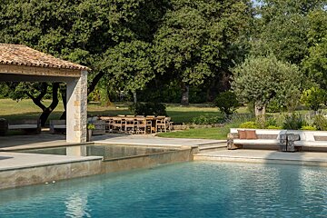 An elegant outdoor space with an infinity pool, covered patio, outdoor dining, lounge seating, and lush green trees.