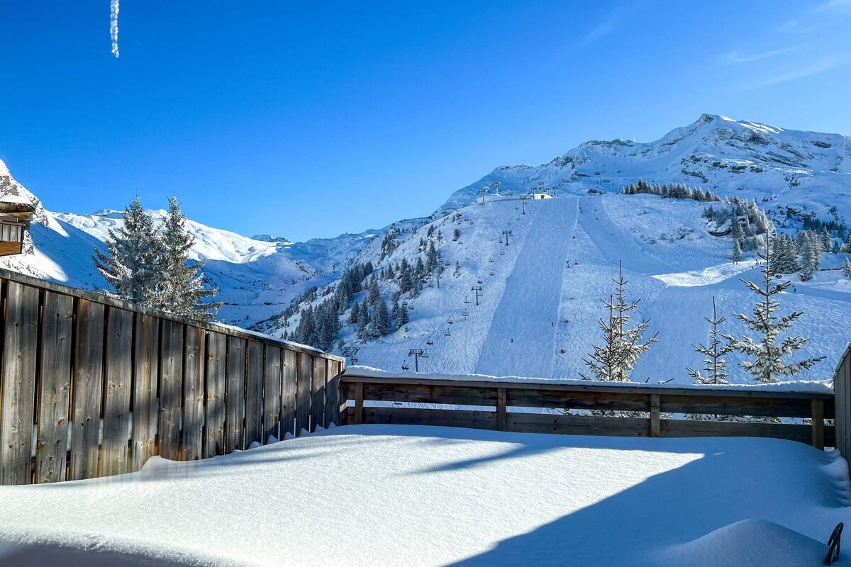 Snow-covered wooden deck overlooking a sunny ski slope and snow-capped mountains under a bright blue sky.