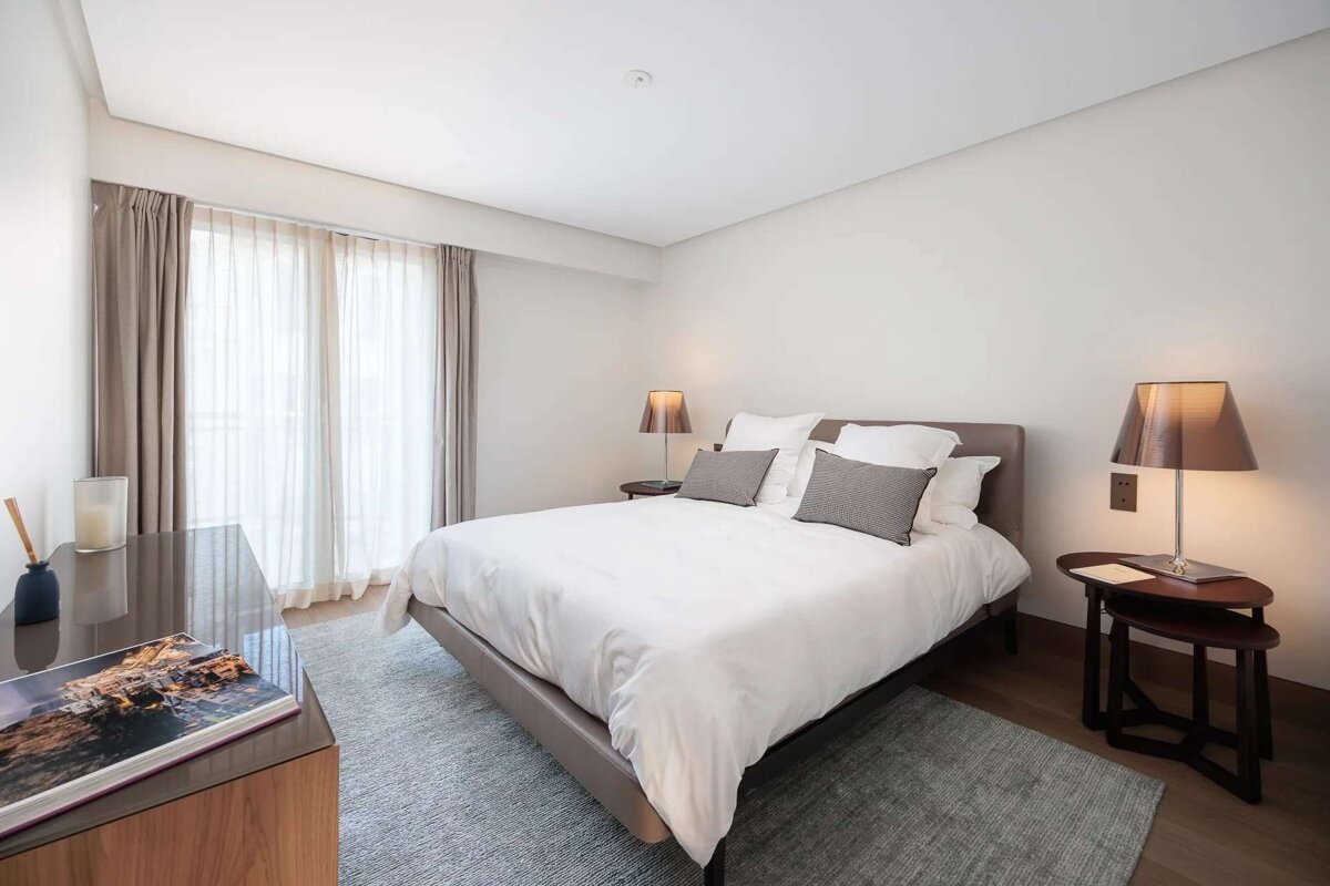 A minimalist bedroom with a large bed, white linens, light gray rug, nightstands with lamps, and sheer curtains over large windows.